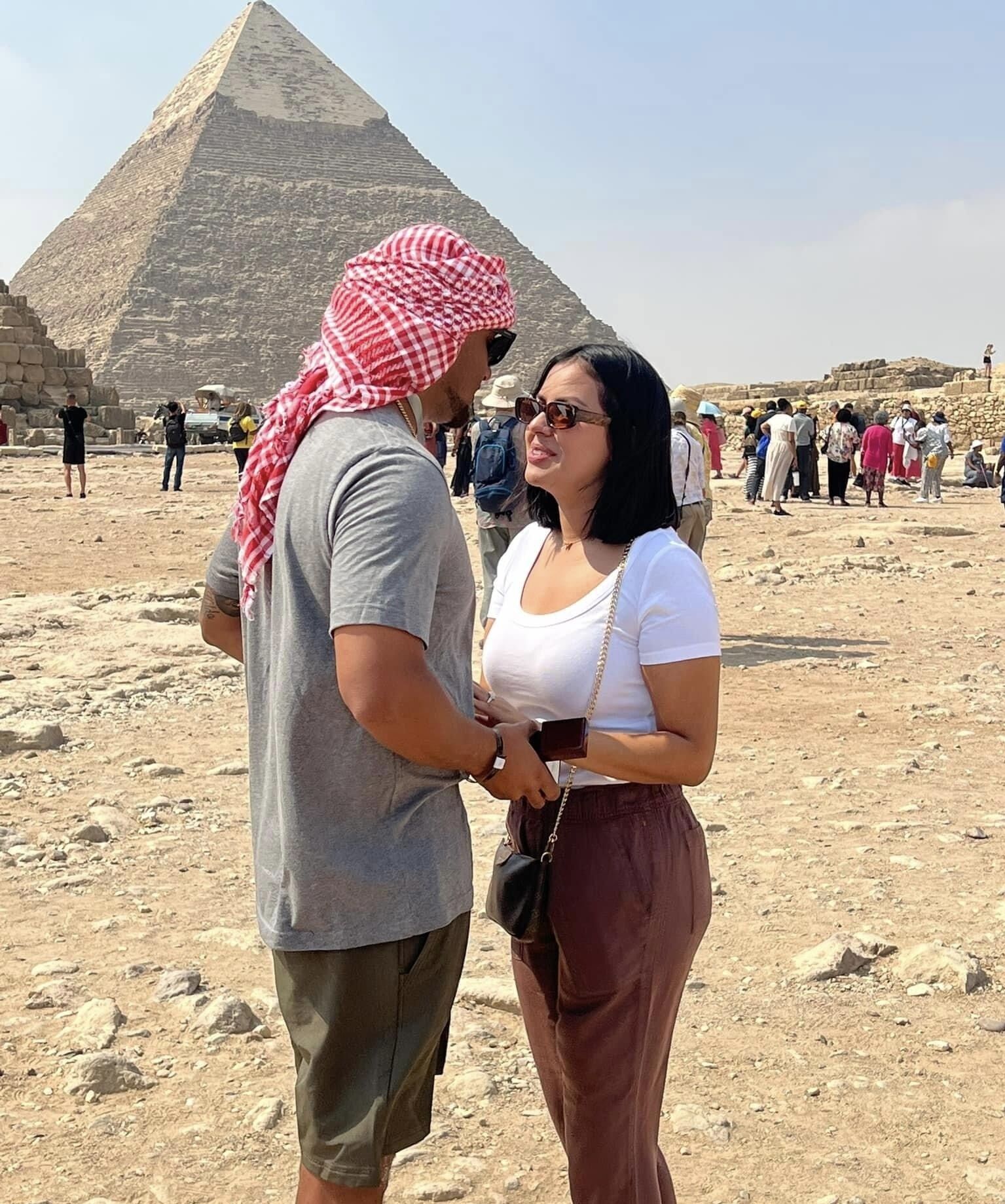Una pareja parada frente a una pirámide, con turistas de fondo.