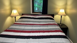 Una cama bien hecha con colcha a rayas, dos lámparas de noche, una ventana con vista a árboles y un interruptor de luz en la pared.