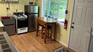Cocina compacta con estufa, horno, microondas y refrigerador. Hay una mesa y sillas de madera, una ventana con vistas y un extintor en la pared.