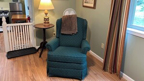 Una sala acogedora con un sillón azul turquesa, una mesa de comedor de madera, una lámpara, un pequeño ventilador y un cuadro en la pared.