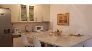 A compact kitchen with a microwave, kettle, and a small refrigerator. There is a dining table with chairs and a bowl of fruit.