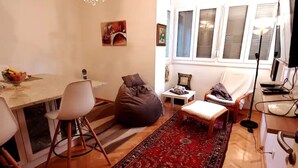 A living room with a wooden floor, a red patterned rug, a white sofa, a bean bag, a small table with a lamp, and a TV on a stand.