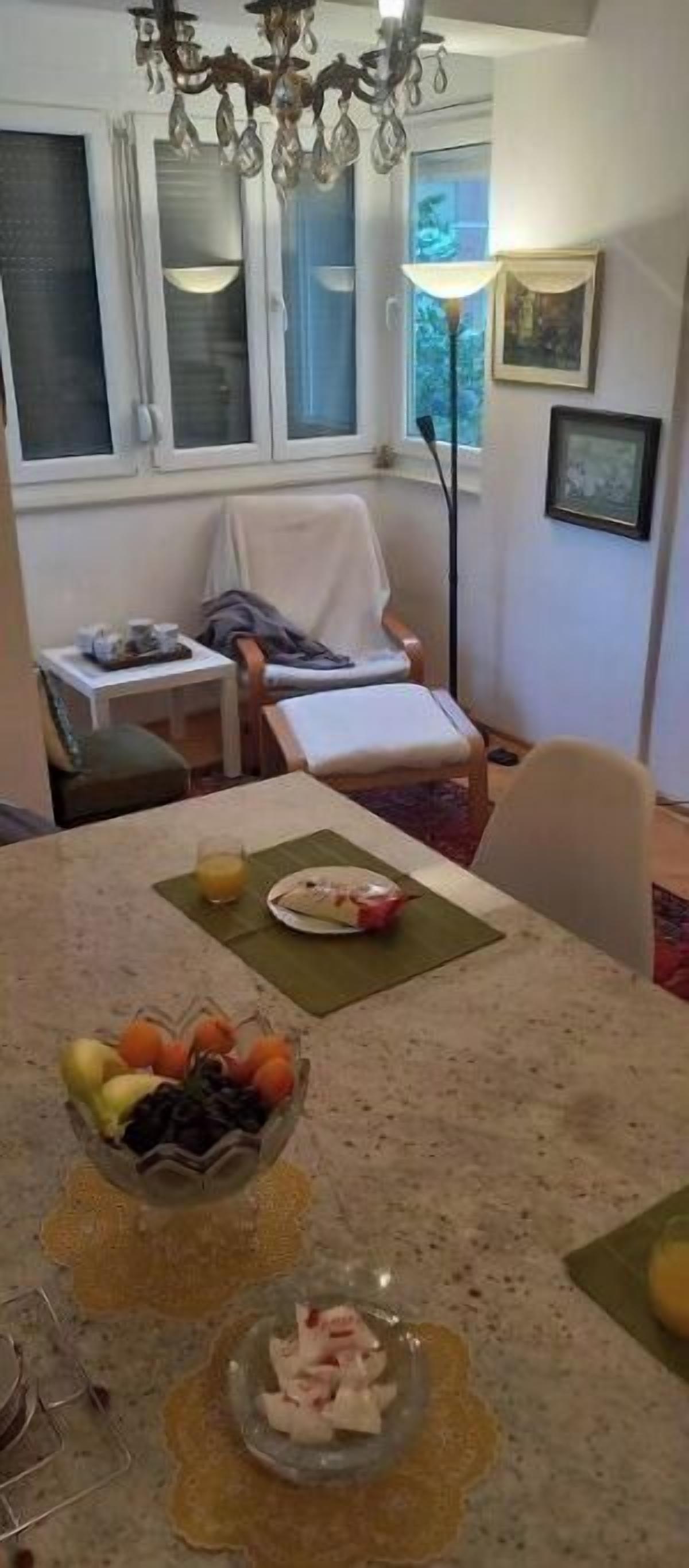 A dining area with a table set for two, a bowl of fruit, and a glass of orange juice. There is a window with blinds, a chair, and a floor lamp.