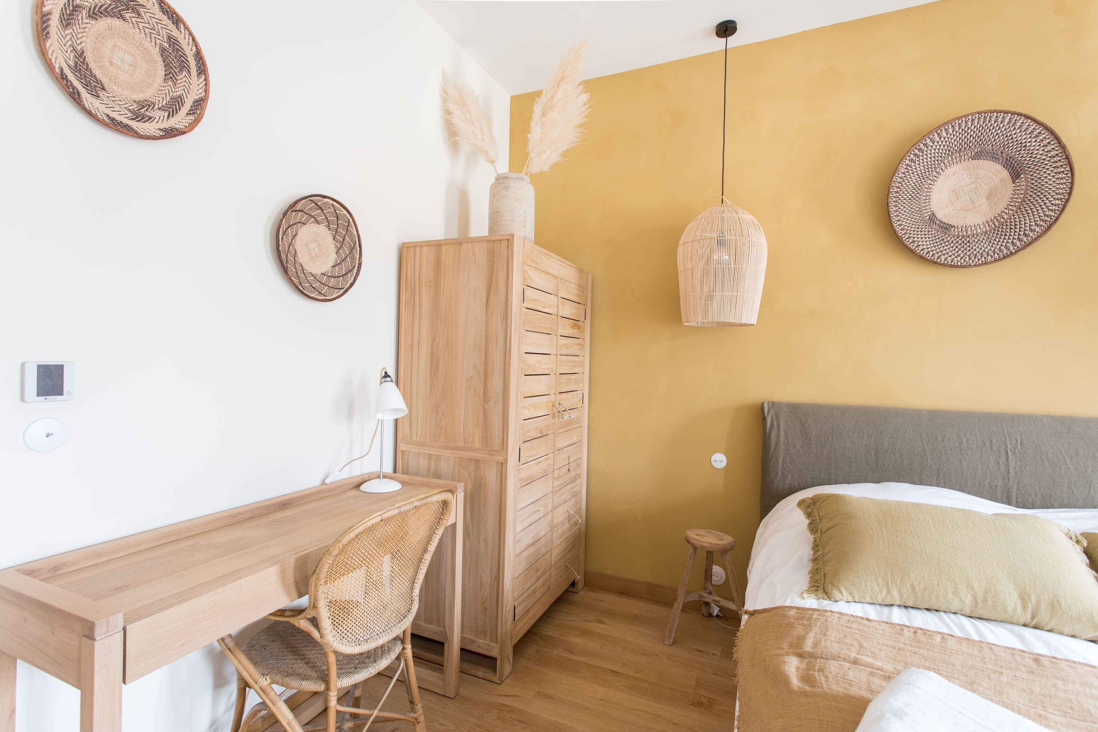 Un dormitorio con un escritorio de madera, una silla, un armario, una cama con cabecera gris y dos lámparas de pared.