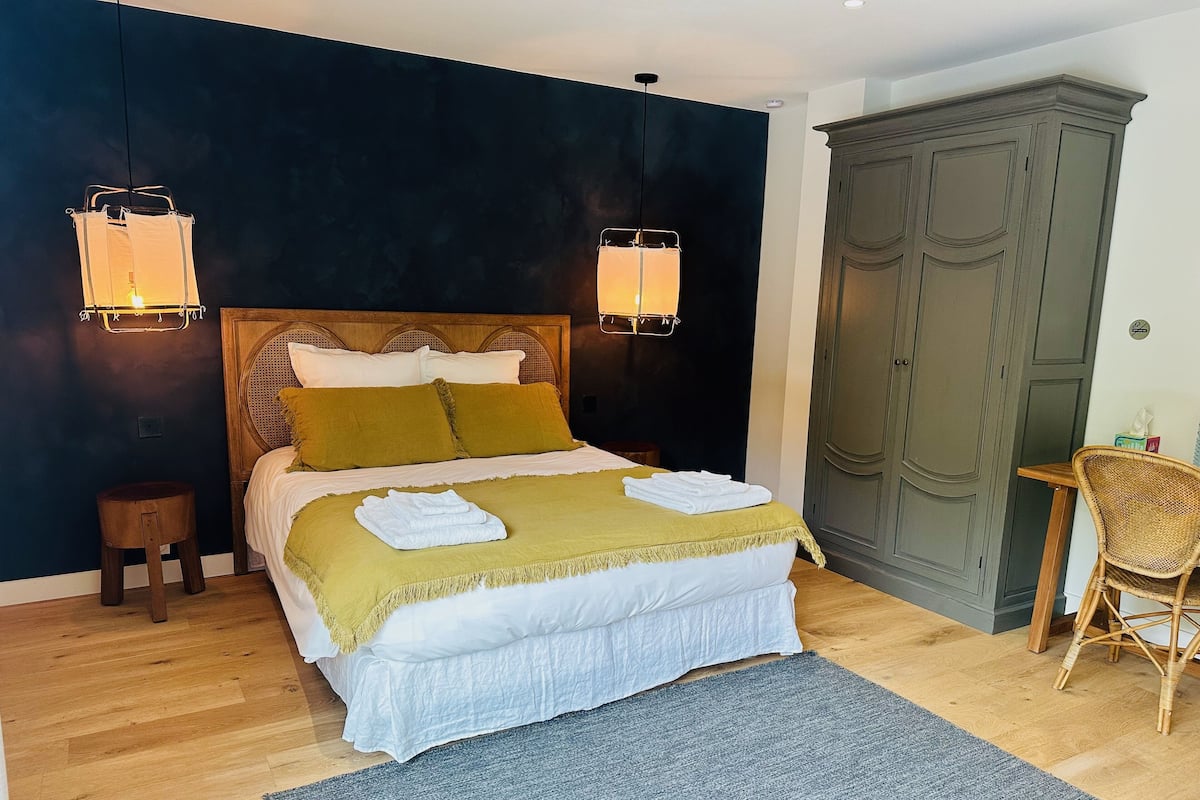 Un letto rifatto con lenzuola bianche, una coperta verde e due cuscini. Si notano due lampade a parete, un comodino in legno, una sedia in vimini e un ampio guardaroba.