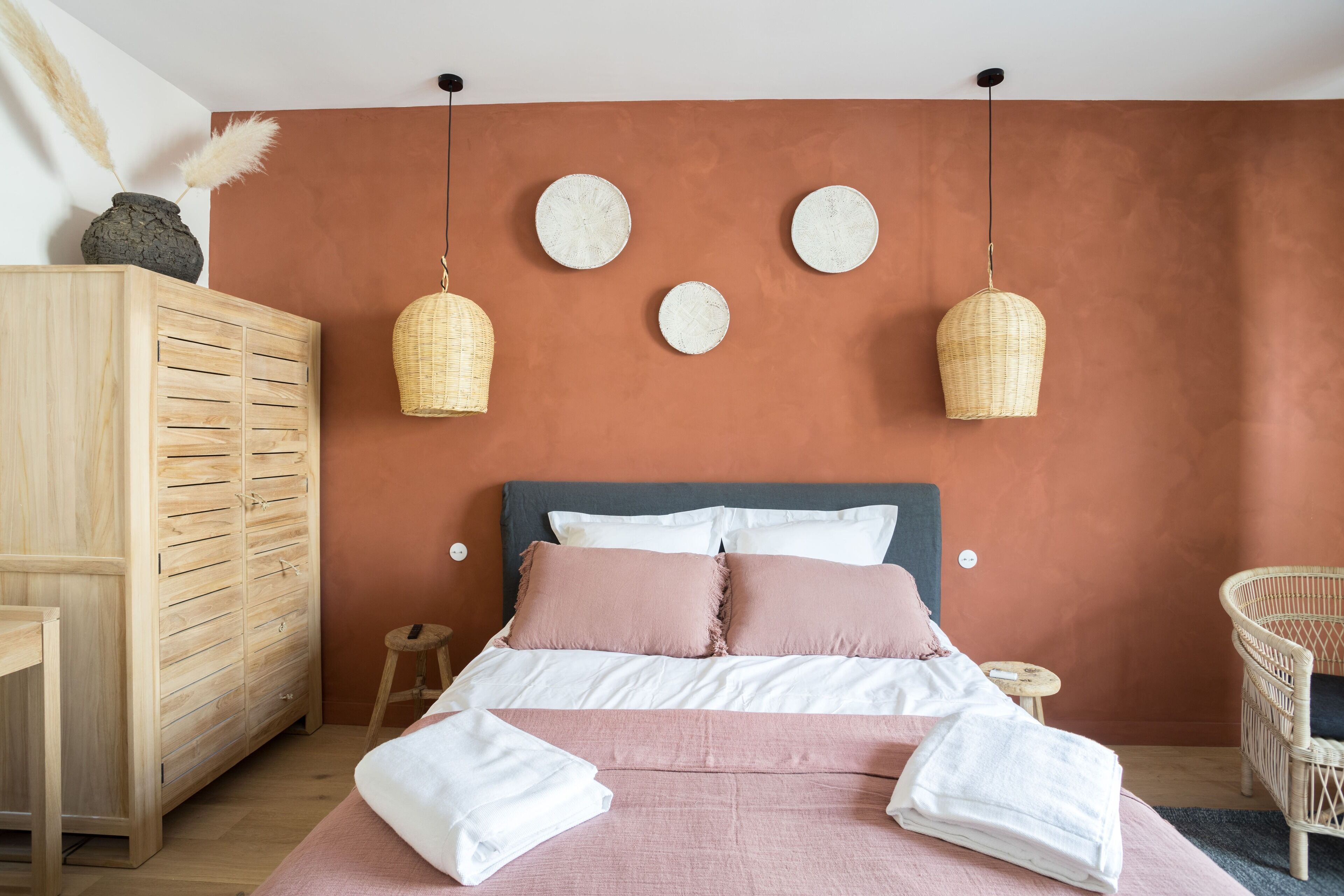 Un dormitorio con cama, armario, taburete pequeño y silla de mimbre.