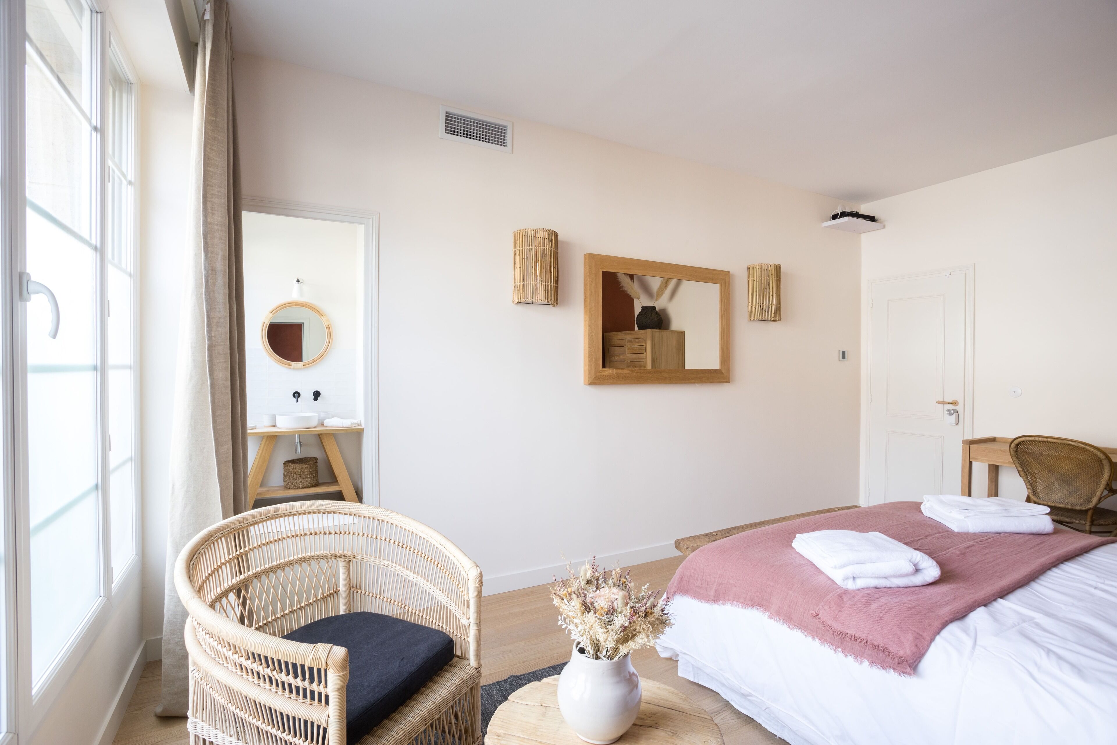A hotel room with a bed, a wicker chair, a small table with a vase, and a mirror on the wall.