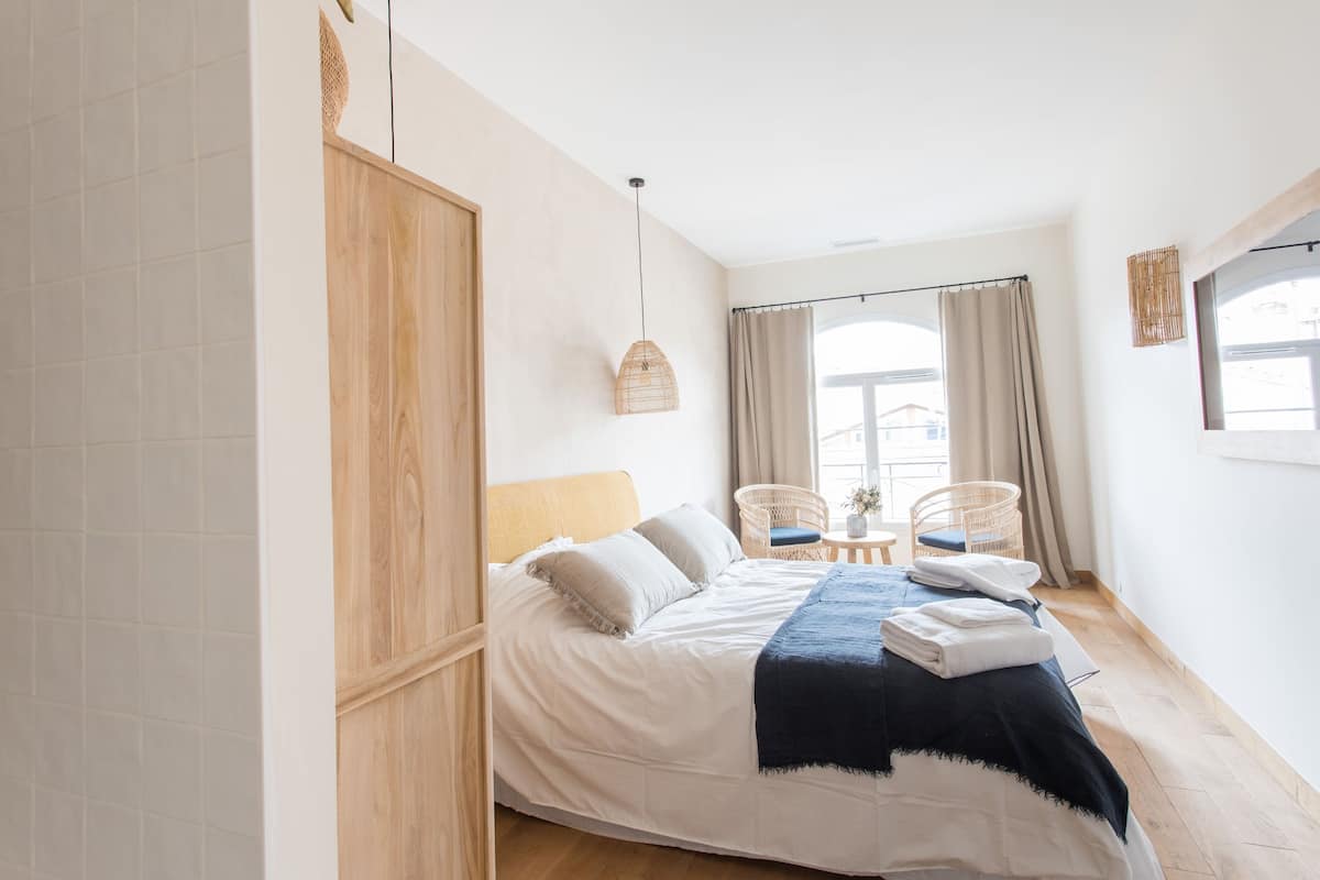 Una camera da letto con un letto, un'armadiatura in legno, una sedia in vimini, un tavolino e una finestra con tende.