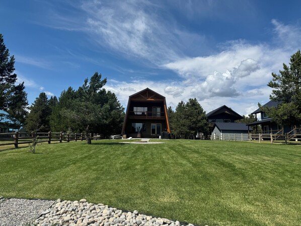 A modern two-story house with a prominent wooden facade, surrounded by a well-maintained lawn and a wooden fence. - Lakefront 4BR/4BA Cabin with Sauna, Hot Tub & Private Dock – Minutes from YNP! (Island Park)