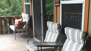 Een overdekt terras met twee gestreepte lounge fauteuils en een kleine tafel, met uitzicht op een bebost gebied.