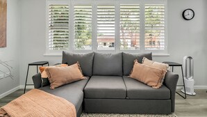 Un salon moderne avec un canapé d’angle gris, deux tables d’appoint, une lampe sur pied et une grande fenêtre avec des stores.