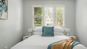 Une chambre à coucher avec un lit, deux tables de chevet, une fenêtre avec des stores et un tableau au mur.