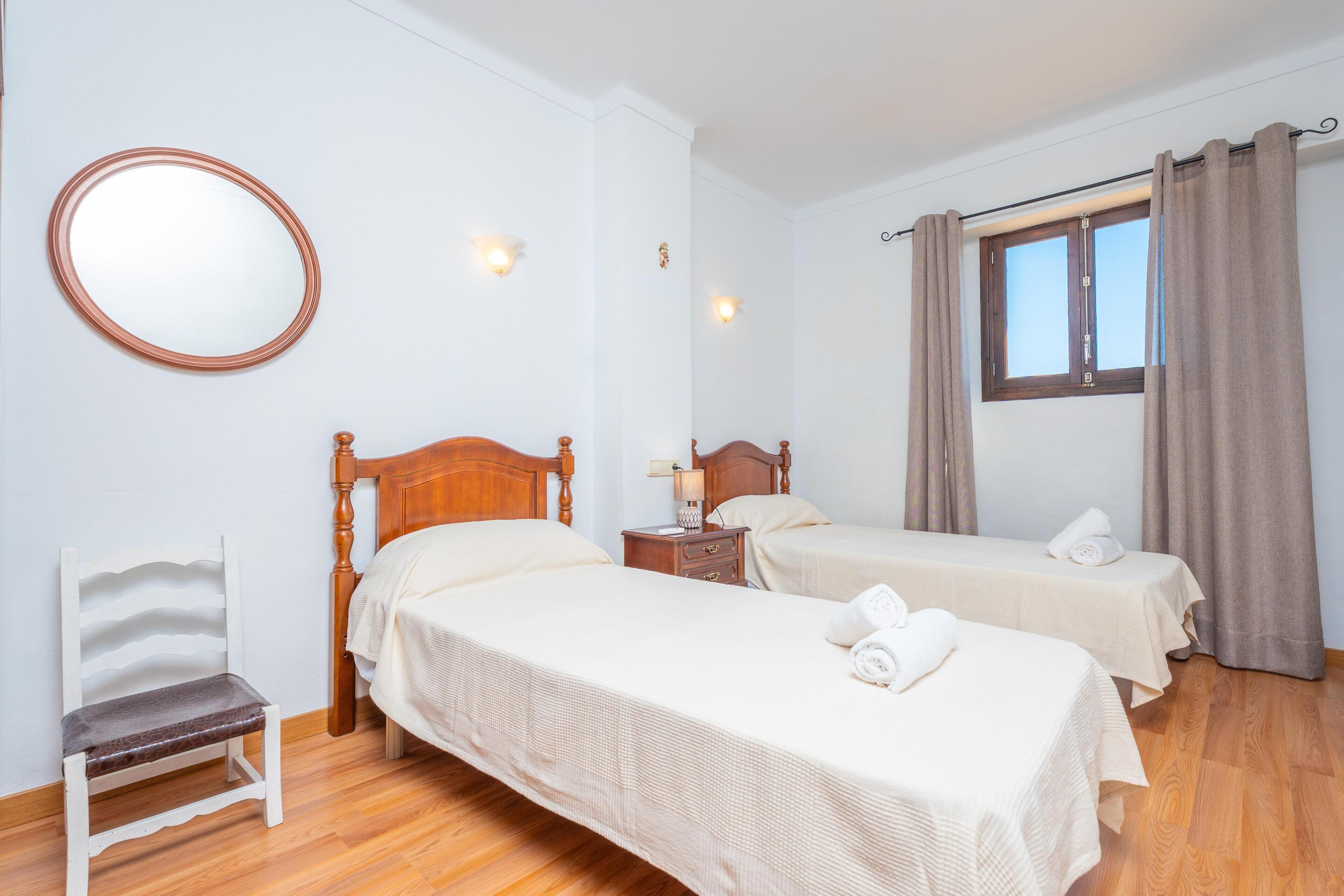 Habitación con dos camas, una silla de madera, un espejo redondo y una ventana con cortinas.