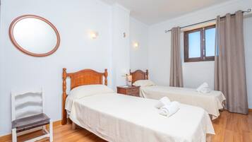 Habitación con dos camas, una silla de madera, un espejo redondo y una ventana con cortinas.