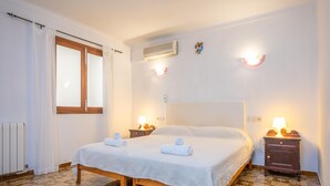 A bedroom with a bed, two bedside tables with lamps, a window with curtains, an air conditioning unit, and a radiator. - CASA CATALINA SAROLA - Village house with a swimming pool in the north of Mallorca (Campanet)