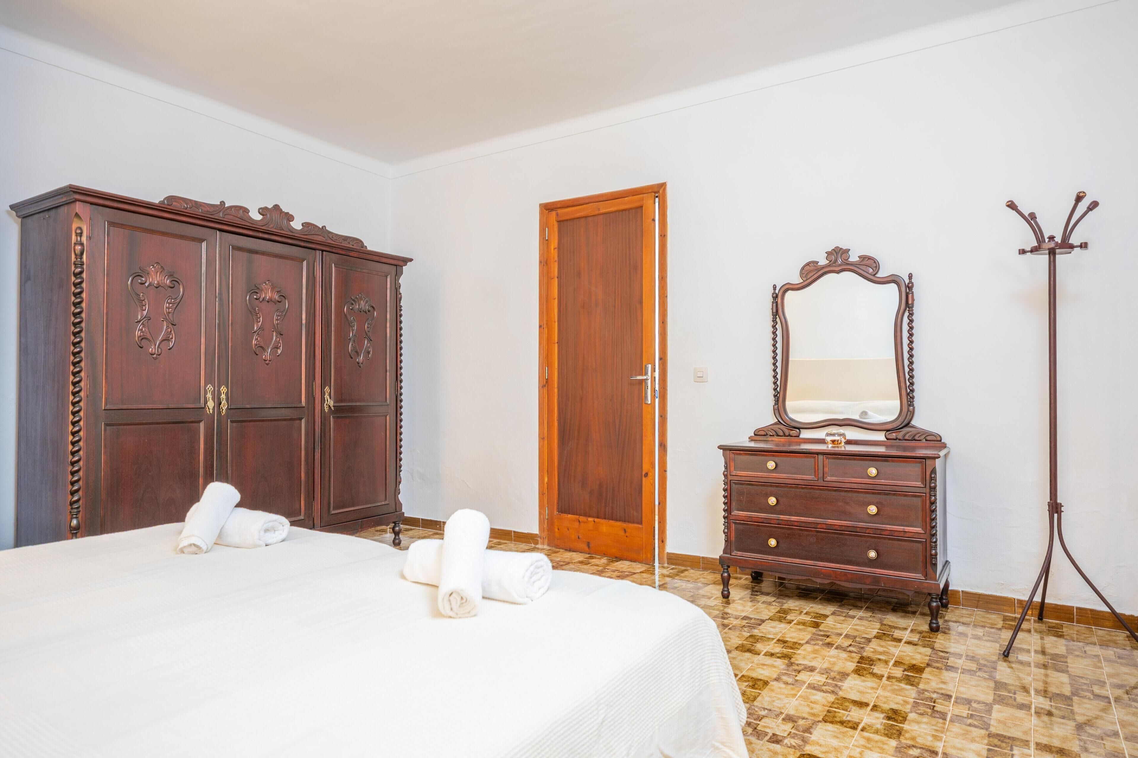 A bedroom with a bed, wooden wardrobe, dresser, mirror, and coat stand.