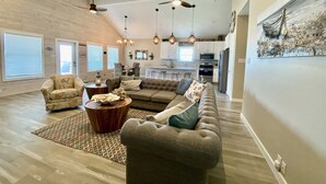 A spacious living room with a large sectional sofa, a patterned armchair, and a coffee table. There are pendant lights and a ceiling fan. The room has wooden flooring and large windows with blinds. - New Listing in Paradise Point Community (Port Aransas)