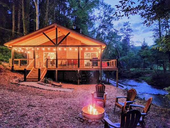 A wooden cabin with a deck, a fire pit, and chairs.