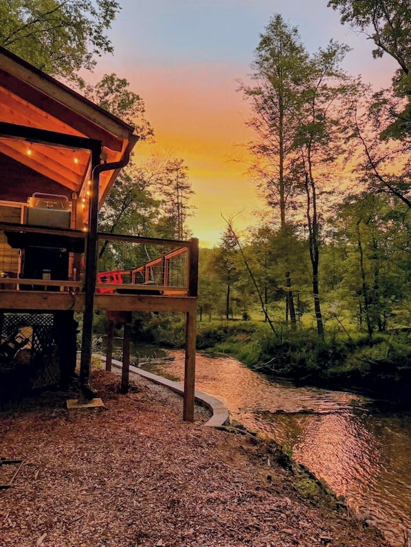 A rustic cabin with a deck overlooking a forested area and a stream.