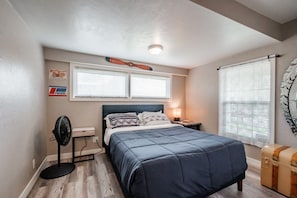Une chambre à coucher comprenant un lit, un ventilateur sur pied, une table de chevet, une lampe, une fenêtre avec des stores et un élément décoratif fixé au mur.
