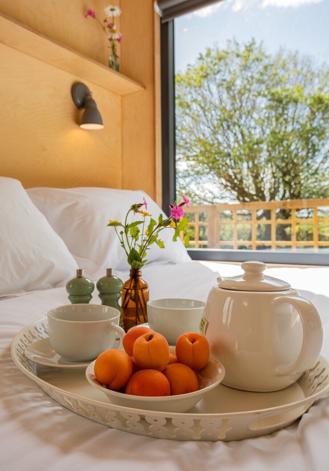 A bed with a tray of apricots, a teapot, and a cup on a hotel room with a view of trees.