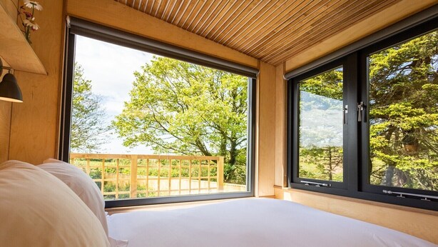 A bedroom with a large window, wooden ceiling, and a view of trees and a wooden deck outside.