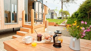A wooden outdoor dining area with a table set for breakfast, including a coffee pot, eggs, and juice. - A secluded, design-led shepherd’s hut for slow, off-grid stays by the coast (Cardigan)