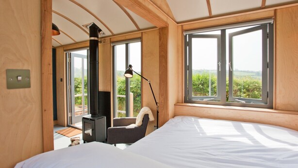 A bedroom with a bed, a chair, a desk, a lamp, and a window with a view of greenery. - A secluded, design-led shepherd’s hut for slow, off-grid stays by the coast (Cardigan)