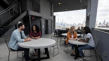 A rooftop terrace with round tables and chairs, a staircase, and a city skyline view.
