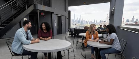 Una terraza en la azotea con mesas y sillas redondas, una escalera y vistas del horizonte urbano.