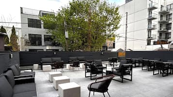 Un espacio al aire libre con mobiliario moderno, un árbol y un edificio al fondo.