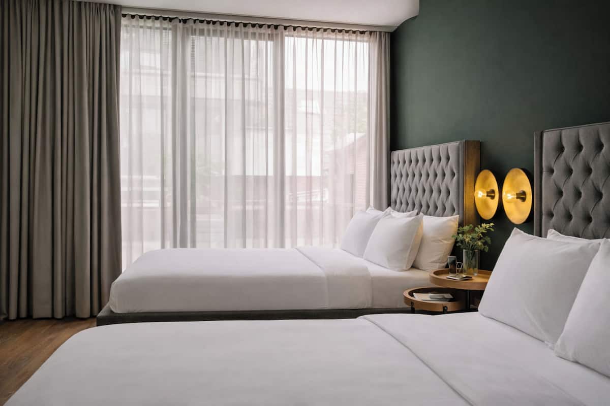 Ein Hotelzimmer mit zwei Betten, einem großen Fenster mit durchscheinenden Vorhängen, einem gepolsterten Kopfteil und einem kleinen Nachttisch mit Lampe und einer Vase.