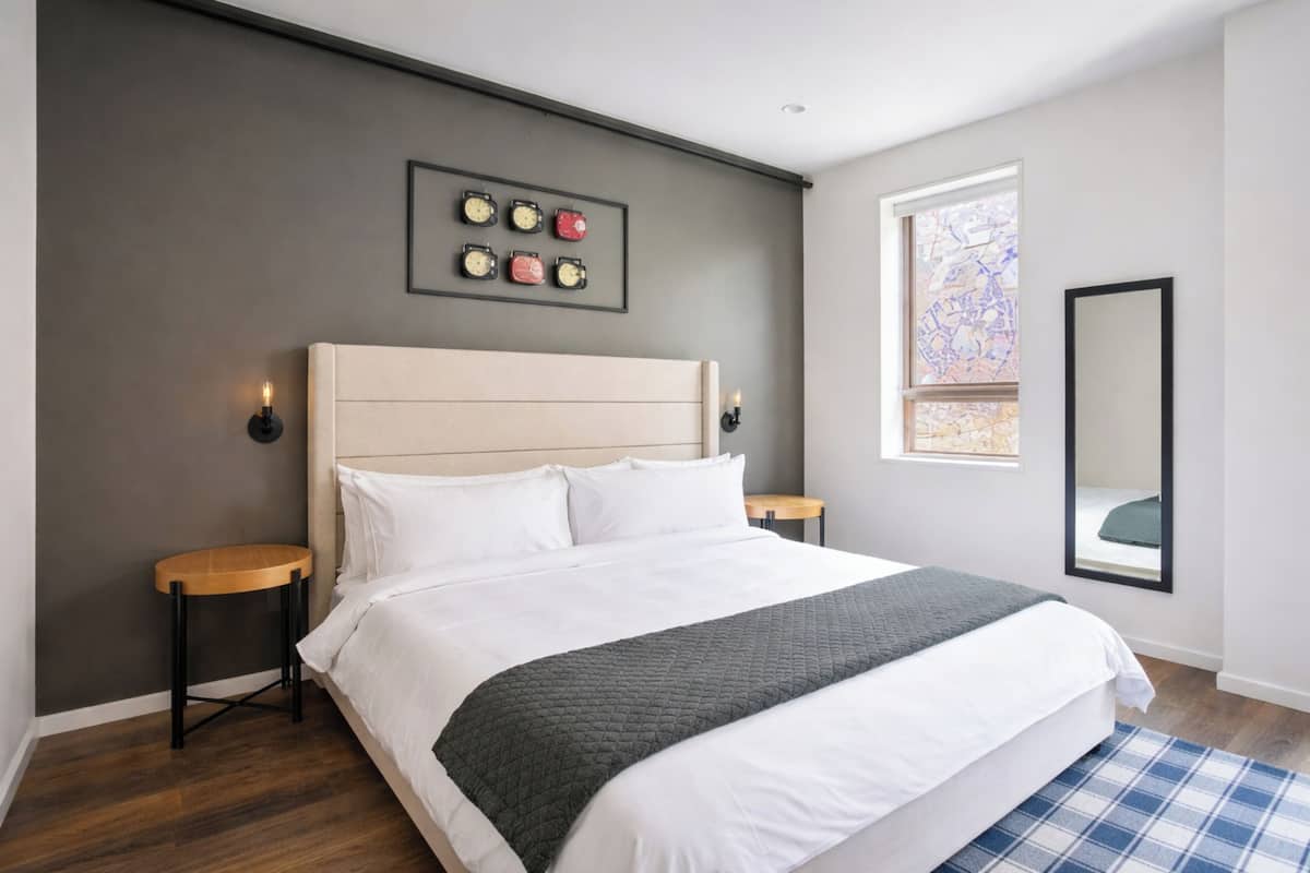 Ein modernes Schlafzimmer mit einem großen Bett, einem Kopfteil, Nachttischen, einer Wanduhr, einem Fenster und einem Spiegel.