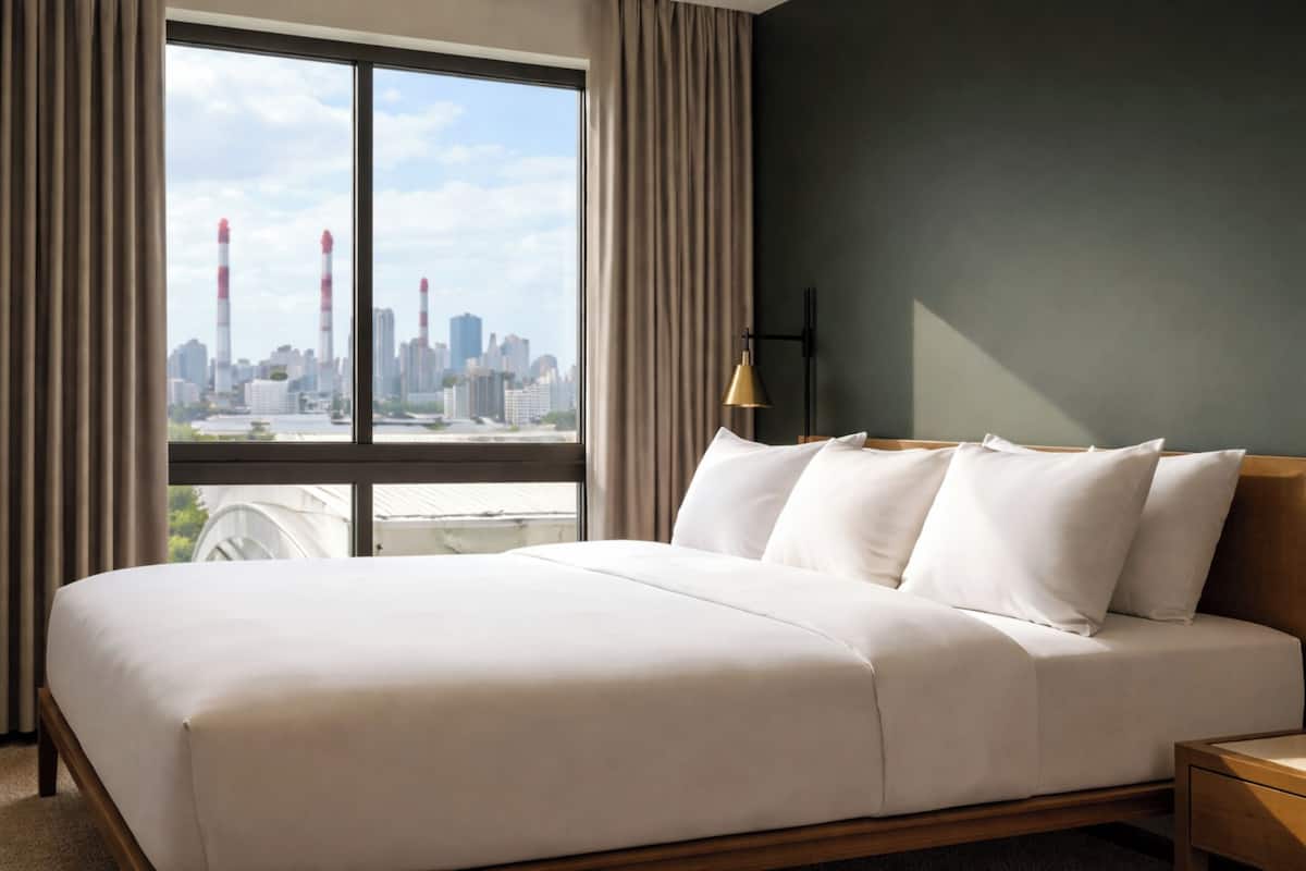 Ein Hotelzimmer mit einem großen Bett, einem Nachttisch, einer Lampe und Blick auf eine Stadtlandschaft durch ein großes Fenster.