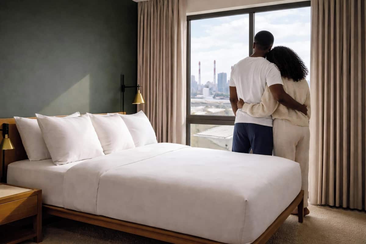 Ein Schlafzimmer mit einem großen Bett, einem Nachttisch, einer Lampe und Blick auf eine Stadtlandschaft durch ein Fenster.