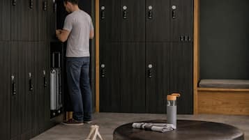 A man standing in a room with dark wooden lockers, a suitcase, a round leather ottoman with a water bottle, and a beige bag on the floor.