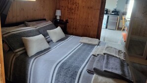 A bedroom with a bed, pillows, a nightstand, a lamp, and a view of a kitchen area.