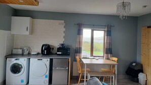 A compact kitchen with a washing machine, a coffee maker, and a refrigerator. There is a dining table with chairs and a view of the outdoors through a window.