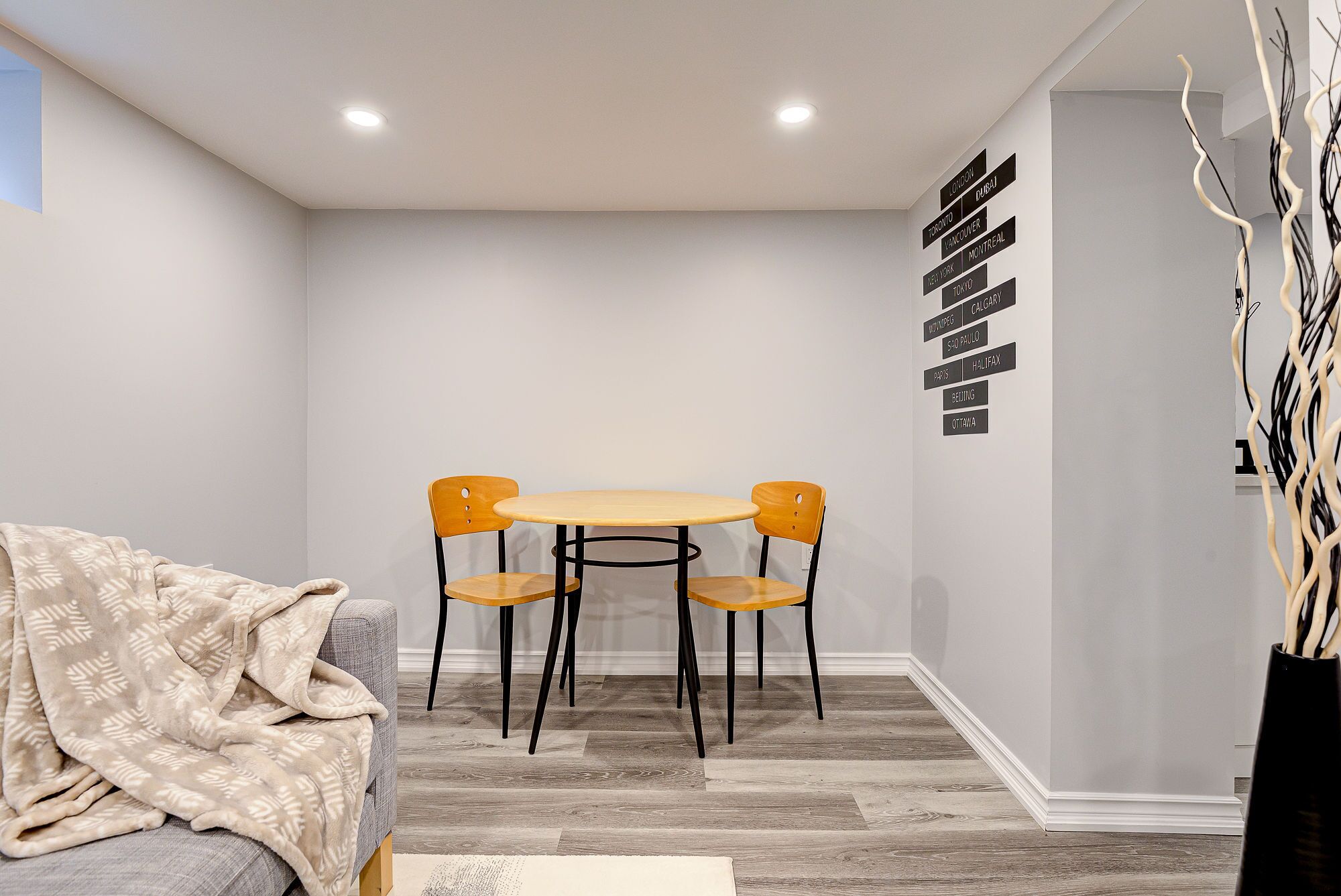 A dining area with a round table and four chairs, a sofa with a blanket, and a wall with city names.