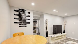 A modern living room with a wooden dining table, a flat-screen TV on a white stand, and a wall with city names. - 1 Bedroom Apartment Downtown - Close to Hospital and University  (Kitchener)