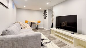 A modern living room with a grey sofa, a TV on a white stand, a dining table with chairs, and a decorative wall piece. - 1 Bedroom Apartment Downtown - Close to Hospital and University  (Kitchener)