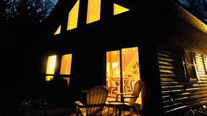 A cabin with a large front window, a dining area with chairs, and a visible staircase leading to the upper level.