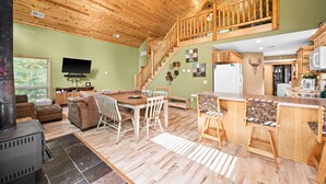 A spacious living area with a wooden ceiling, a fireplace, a dining table with chairs, a kitchen area with a refrigerator, and a staircase leading to an upper level.