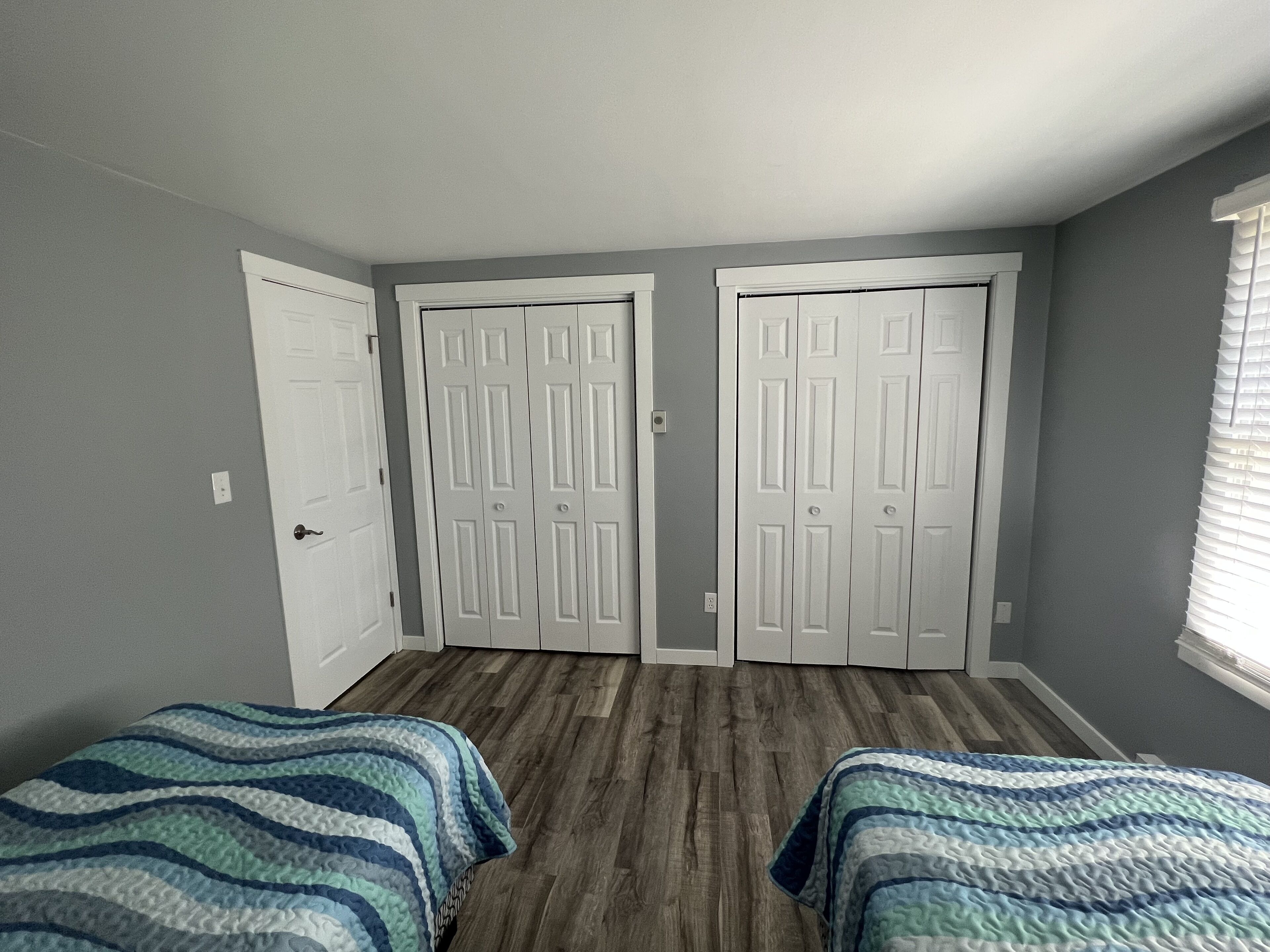 Ein Zimmer mit zwei Betten, grauen Wänden, Holzböden und weißen Türen.