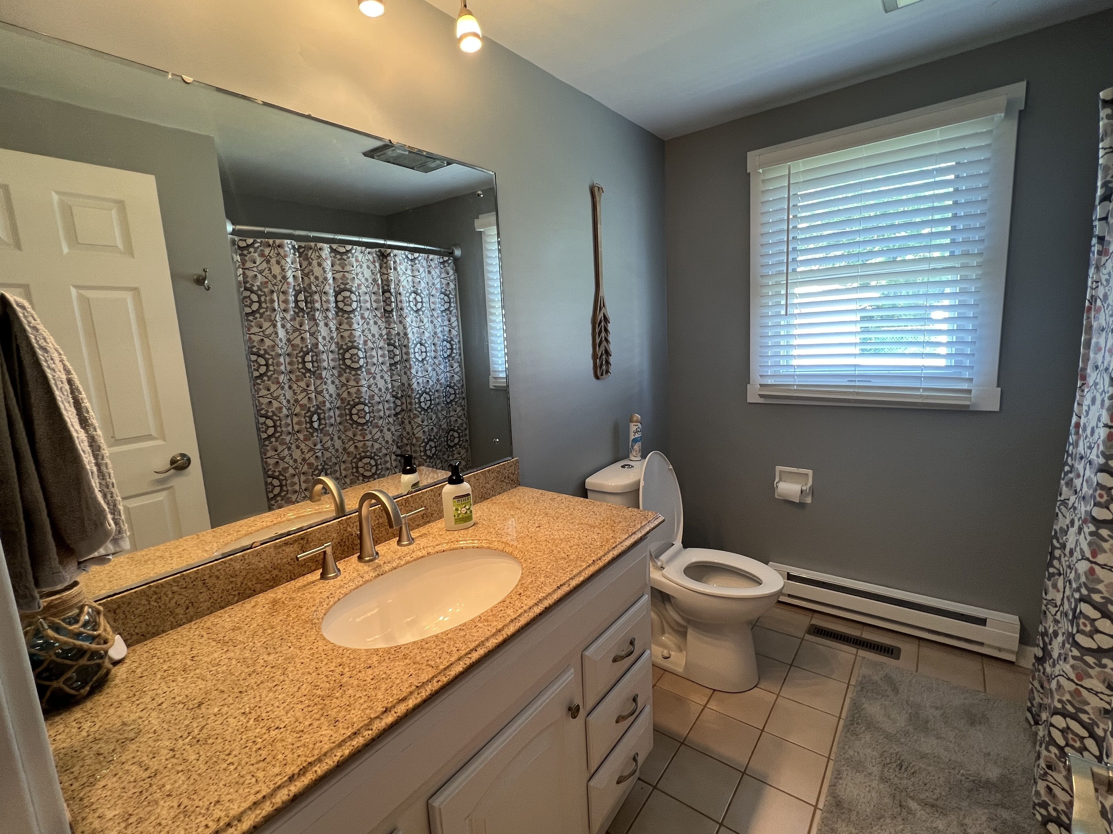 A bathroom with a granite countertop sink, a toilet, a shower curtain, and a window with blinds.