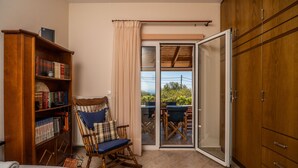Quarto com estante de madeira, poltrona de balanço, mesa de jantar com cadeiras e porta de vidro corrediça que dá para uma área externa.