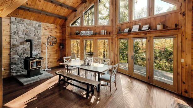 A cozy cabin interior with a stone fireplace, wooden walls, large windows, and a dining area. - Log Cabin with living room fireplace and hot tub (Amberson)