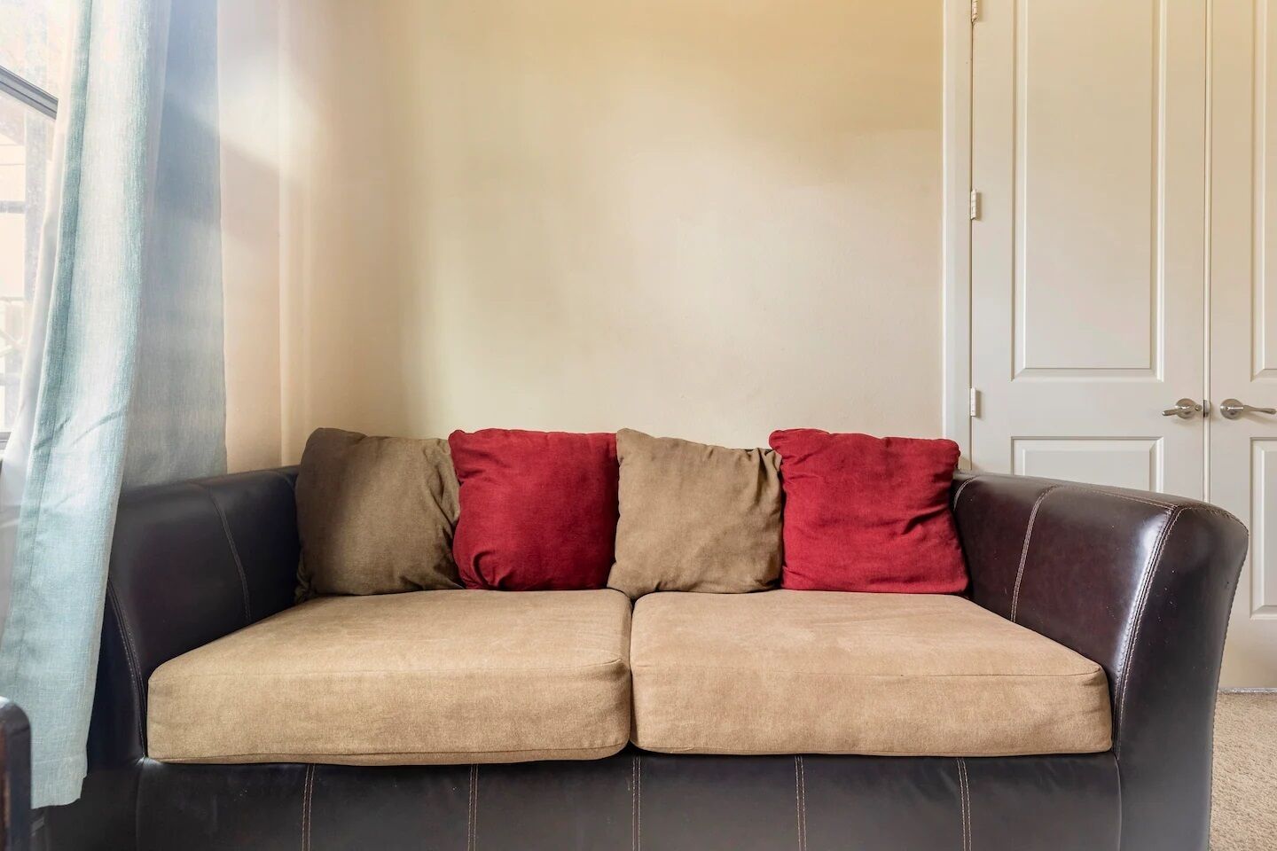 A beige sofa with two red cushions and a dark brown leather armrest.