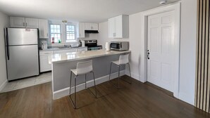 A modern kitchen with white cabinets, a stainless steel refrigerator, a microwave, and a stove. There are two bar stools and a window providing natural light. - Charming cottage in Hyannis walk to downtown, beach, Harbor, pet friendly fence (Barnstable)