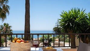 Una mesa con un frutero, dos copas de vino y un cuenco de papas fritas en un balcón con vistas al mar.